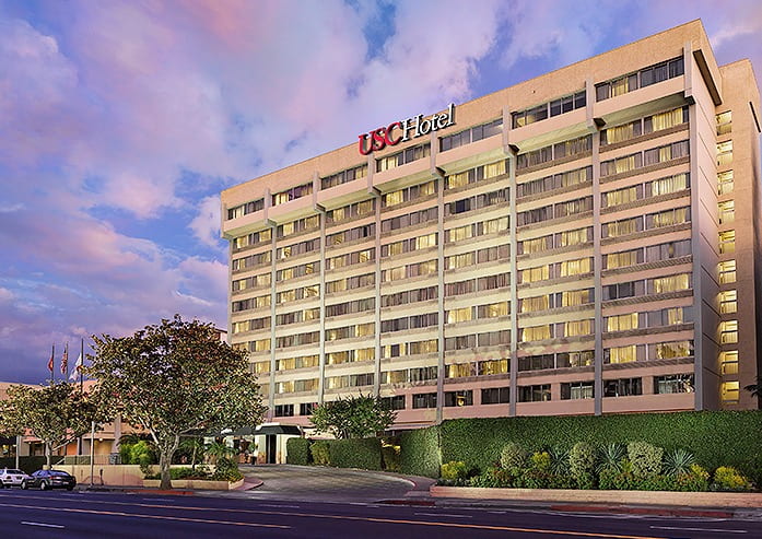 The exterior of USC Hotel during sunset. The sky is pink, purple, and blue. The hotel is a large white building with many windows and a sign on top that reads USC Hotel.