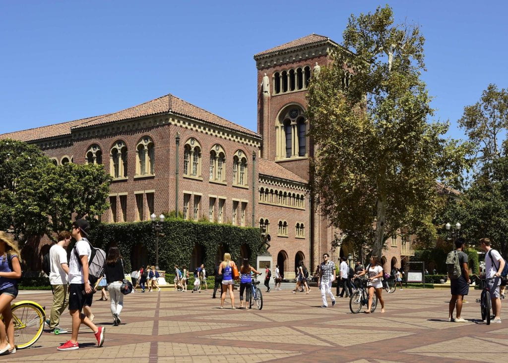 Students walk and ride bikes on Trousdale Parkway on a sunny afternoon. Bovard Auditorium, a large brick building, sits in the background, and the statue of Tommy Trojan is in the center.